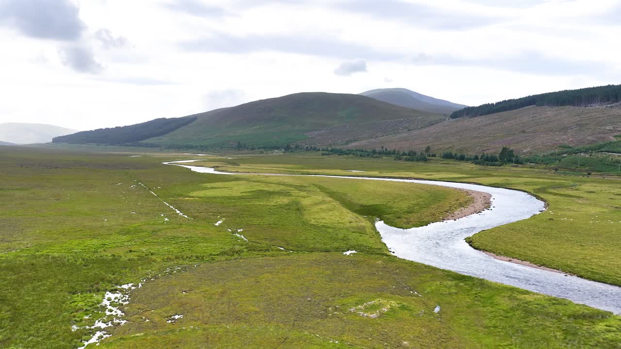 Drone glides above tranquil river, green meadows, and rolling hills under soft daylight