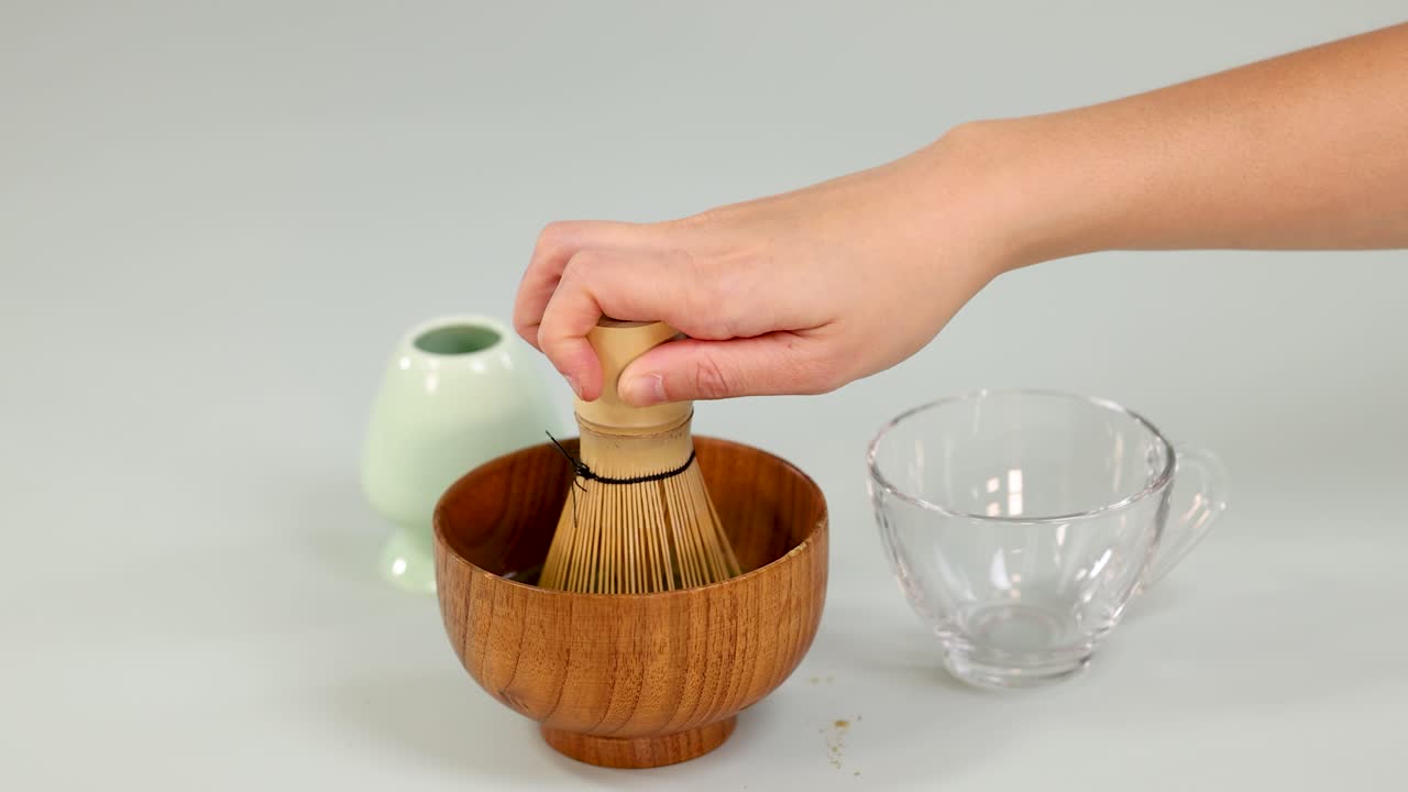 Hand whisks matcha green tea in wooden bowl under soft lighting, static overhead camera angle