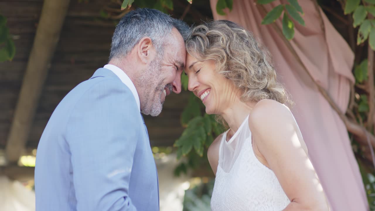 Coppia di sposi che si tengono per mano, appoggiando le fronti sotto un pergolato, in abito, con anelli visibili, festeggiando.