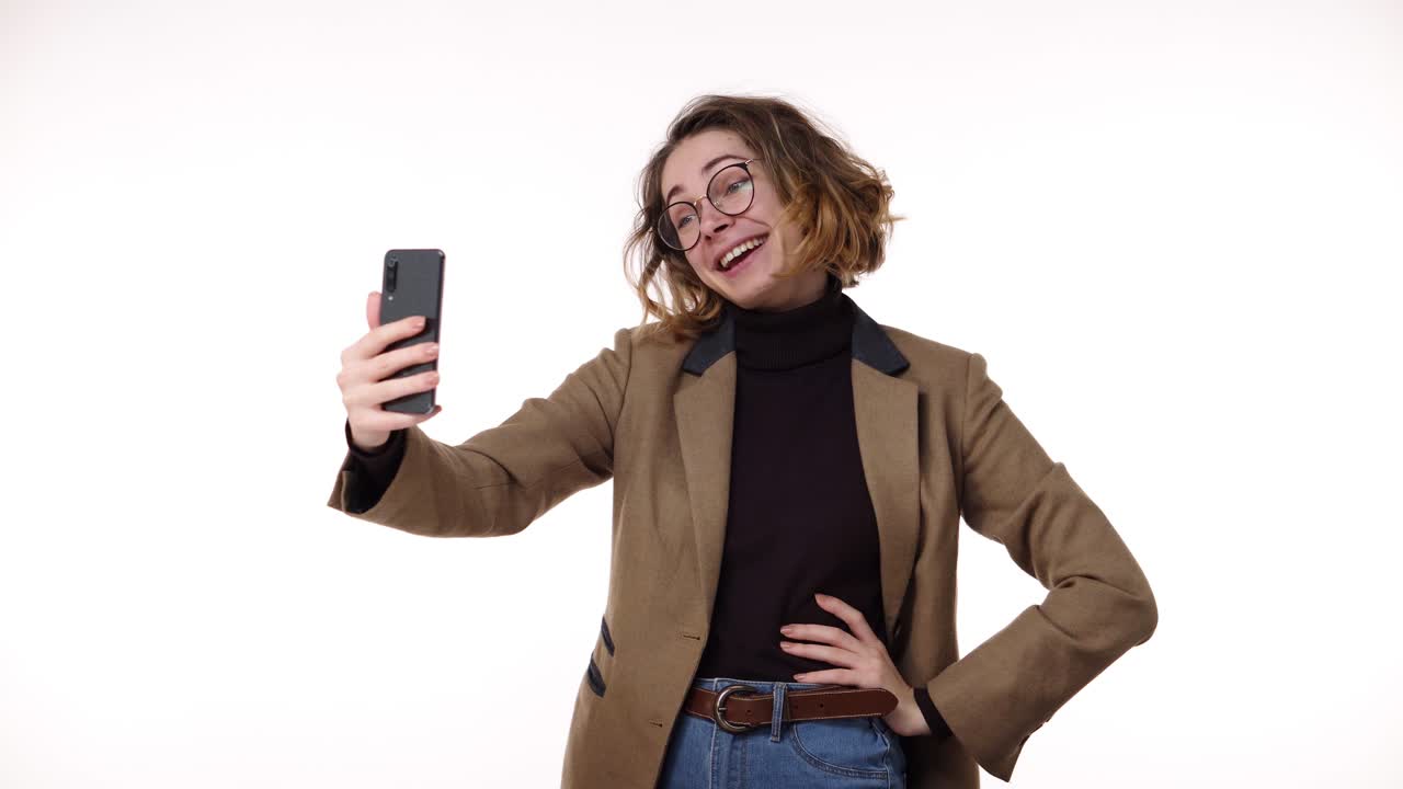 Charming trendy young woman with curly short hair having video chat through phone and laughing, talking standing isolated on white background. Wearing brown jacket and eyeglasses