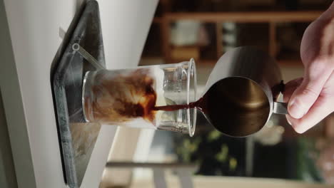 video vertical barista casero haciendo café con leche helado vertiendo espresso en una taza de vidrio que contiene leche con pajita de vidrio en cámara lenta de cerca-1