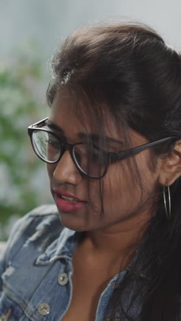 indian woman with ponytail puts on black-rimmed glasses to read book on blurred background. young female student enjoys studying at home closeup