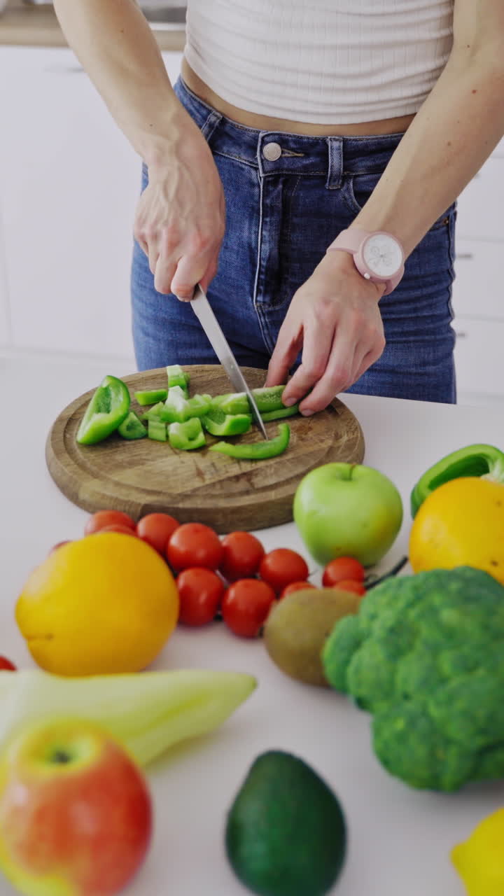 Organic vegetables and fruit on the table. Woman is cutting green pepper on a wooden board to prepare healthy food. Dieting concept. Vertical video