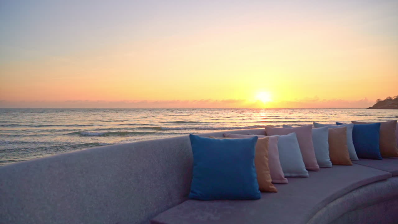Brightly colored pillows are lined up on an empty seaside couch, in the background, is a dynamic sunset floating along the horizon. copy space