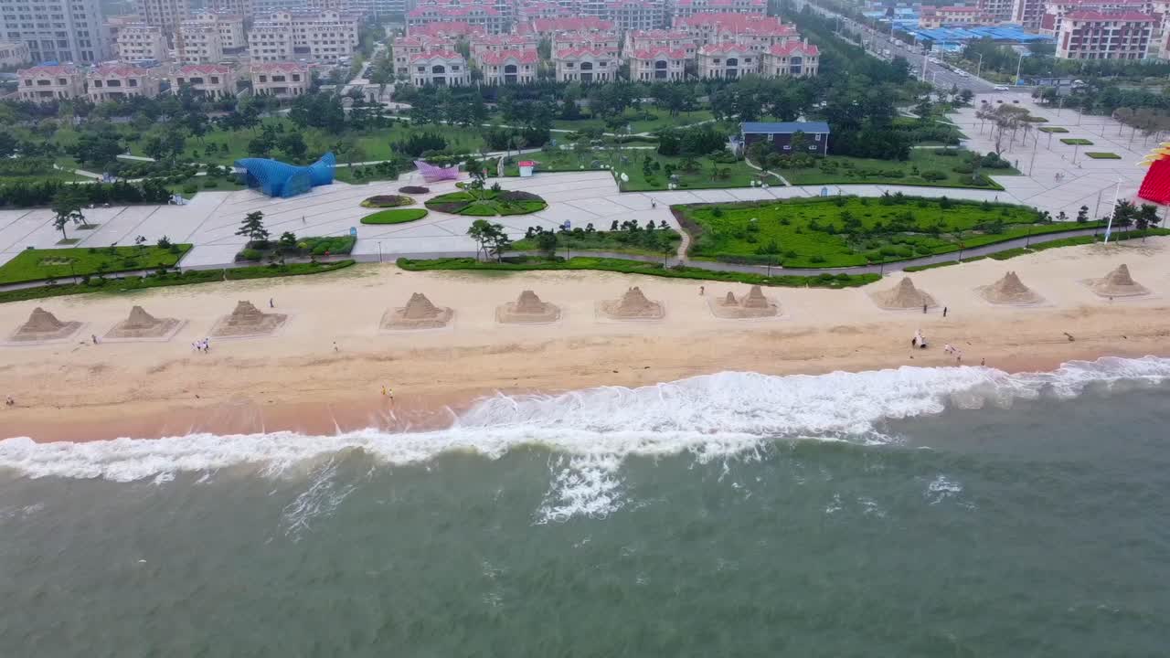 Sliding aerial cinematic view of sand castle sculptures on beautiful beach with crashing waves and park, cityscape background
