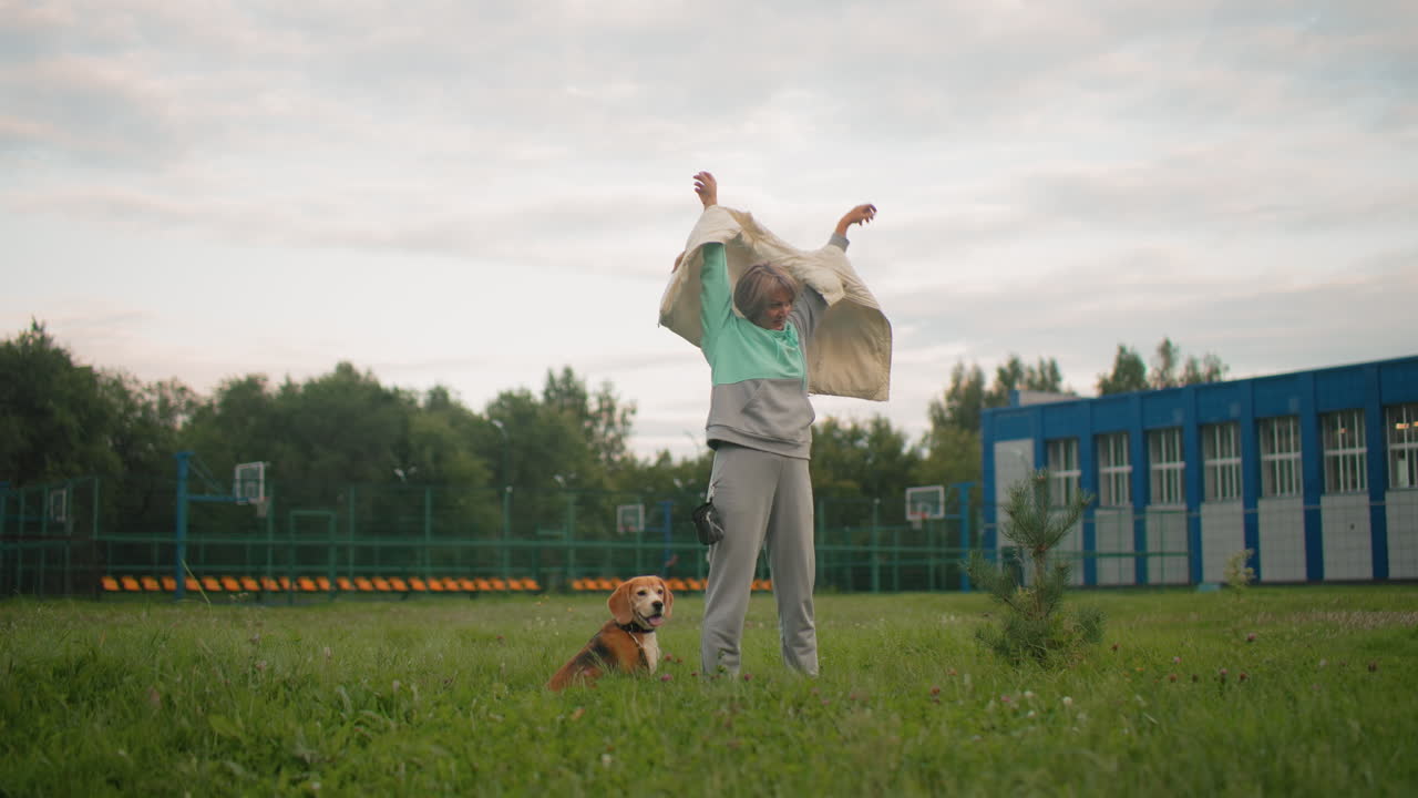 Dog owner zips her rain coat while standing on football field, preparing for dog training session, her dog is visible in the background, actively moving around on grassy field, ready for training