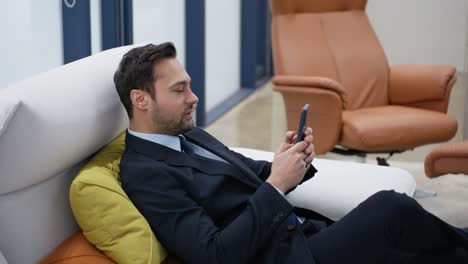 smiling businessman using a mobile phone, typing an email, gaming on smartphone while relaxing reclines on couch in a positive mood with a satisfactory smile at a luxury executive modern office.