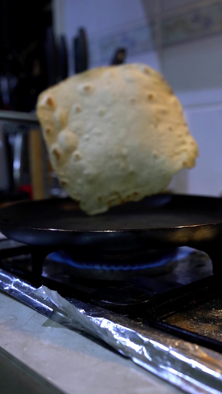 Nærbilde av hånd som snur chapati-deig på svart stekepanne over blå gassflamme. Kjøkkenbakgrunn med knivblokk, uskarpe redskaper og hvit benkeplate. Demonstrasjon av matlaging av hjemmelaget indisk flatbrød