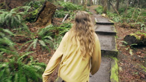 a young woman wearing a bright yellow jacket walks over a small bridge in a green mossy forest, tracking shot from behind