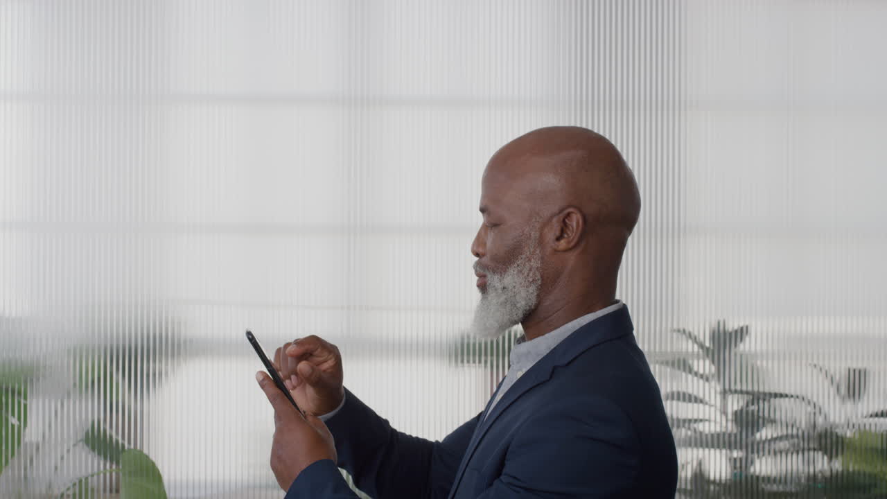 ritratto uomo d'affari afroamericano maturo che usa uno smartphone che si gode di messaggi di testo, di navigazione, di invio di e-mail, di sms sul cellulare in ufficio, vista laterale in slow motion