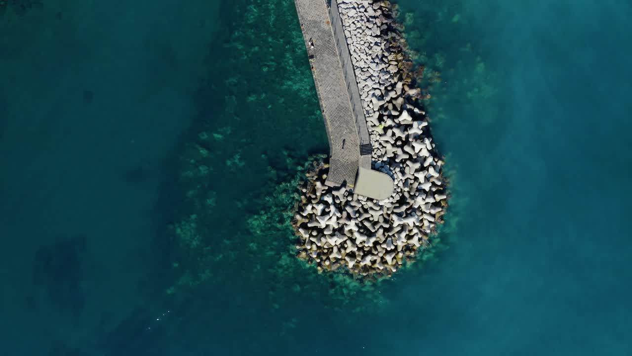 Veduta aerea di persone che camminano su un molo frangiflutti costiero con barriere frangiflutti a tetrapodi in un vibrante paesaggio marino turchese. Litorale della Costiera Amalfitana