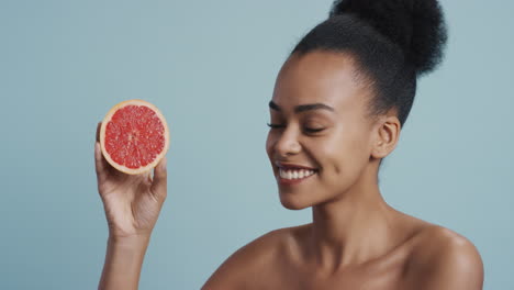 Porträt-Einer-Attraktiven-Jungen-Afroamerikanerin-Mit-Grapefruit,-Die-Lächelnd-Die-Natürliche,-Gesunde-Hautpflegeessenz-Genießt.-Schöne-Frau-Mit-Perfektem-Teint-Auf-Blauem-Hintergrund