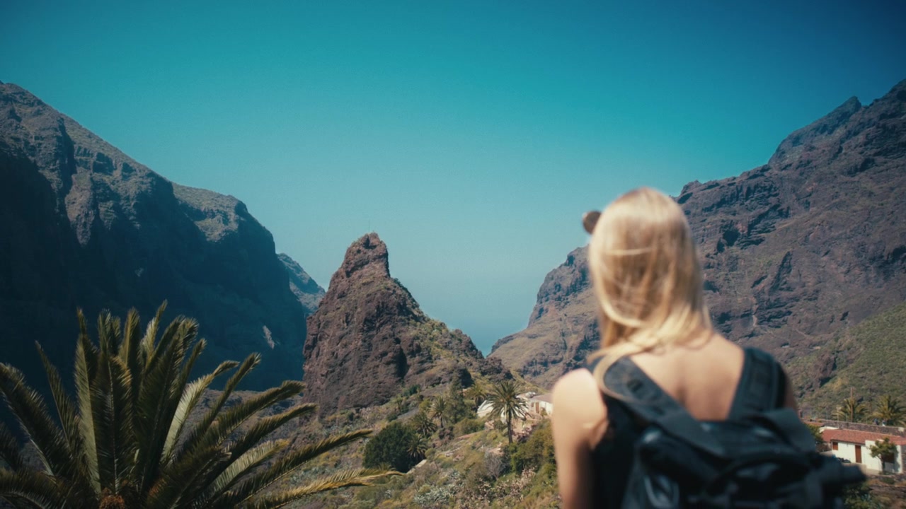 Vista posteriore di una viaggiatrice bionda con zaino che osserva le spettacolari montagne vulcaniche e l'iconica formazione rocciosa della Valle di Masca in una giornata di sole a Tenerife, Isole Canarie