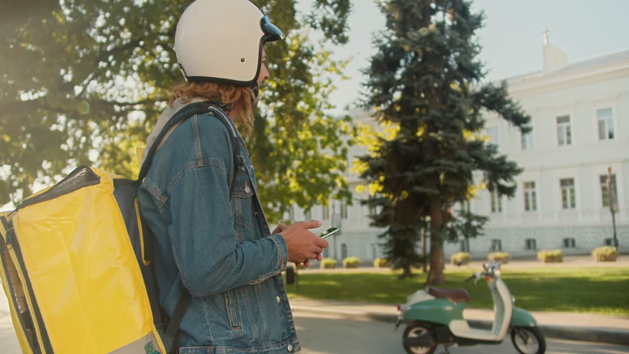 Side view of a guy courier with curly hair in a denim jacket and a white helmet for a moped walks along with a large yellow bag on his shoulders, sits on his green moped and goes to deliver an order in a sunny city park in the summer