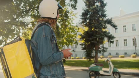 Side-view-of-a-guy-courier-with-curly-hair-in-a-denim-jacket-and-a-white-helmet-for-a-moped-walks-along-with-a-large-yellow-bag-on-his-shoulders,-sits-on-his-green-moped-and-goes-to-deliver-an-order-in-a-sunny-city-park-in-the-summer