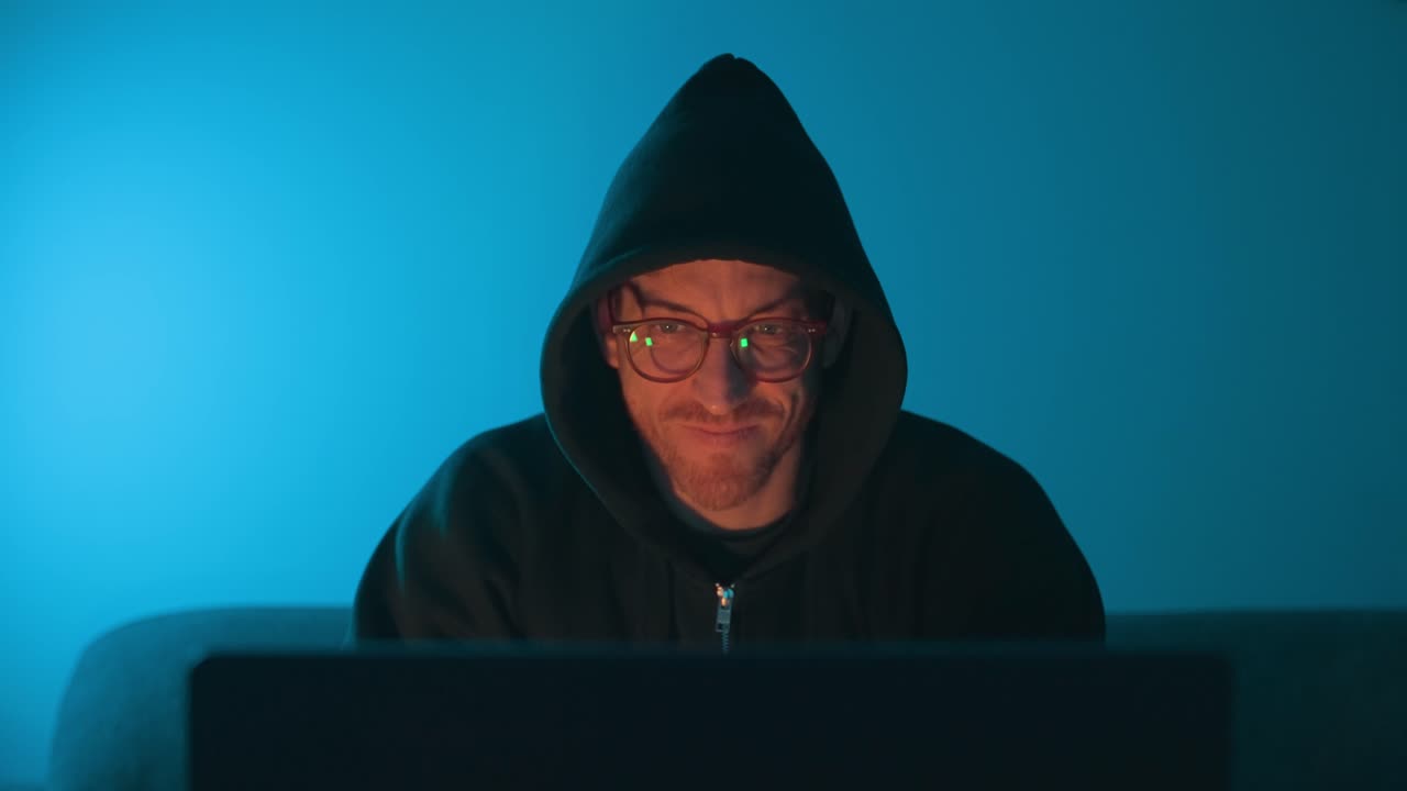 Hacker in hoodie smiling at laptop with blue light, cybersecurity threat concept. Hooded man using laptop, smiling at the screen. Cybersecurity, hacking, cybercrime, data breach, malware. tech