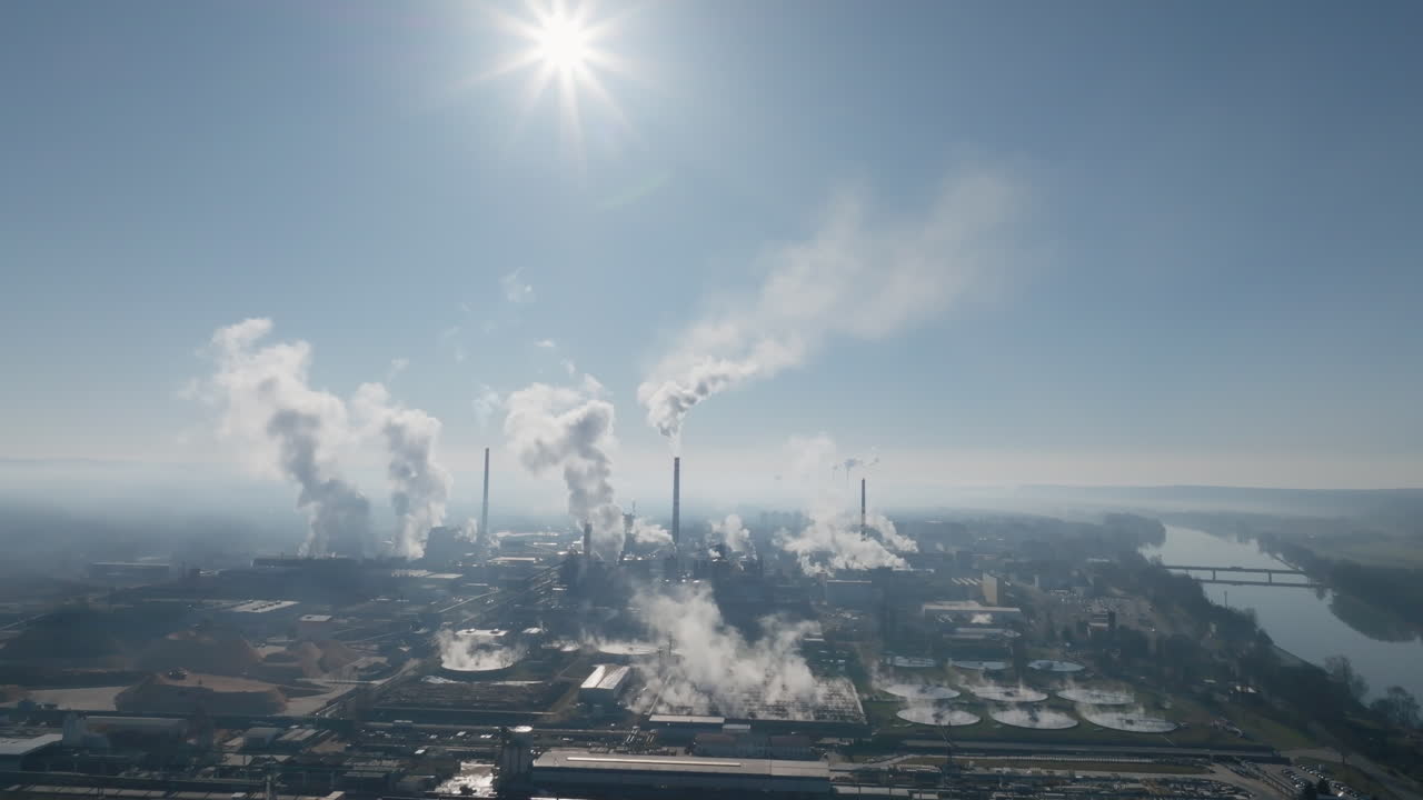 Veduta con drone illuminata dal sole della cartiera di Štětí con imponenti ciminiere e grandi pennacchi di vapore che si innalzano in un cielo azzurro limpido, creando un'atmosfera drammatica in controluce.