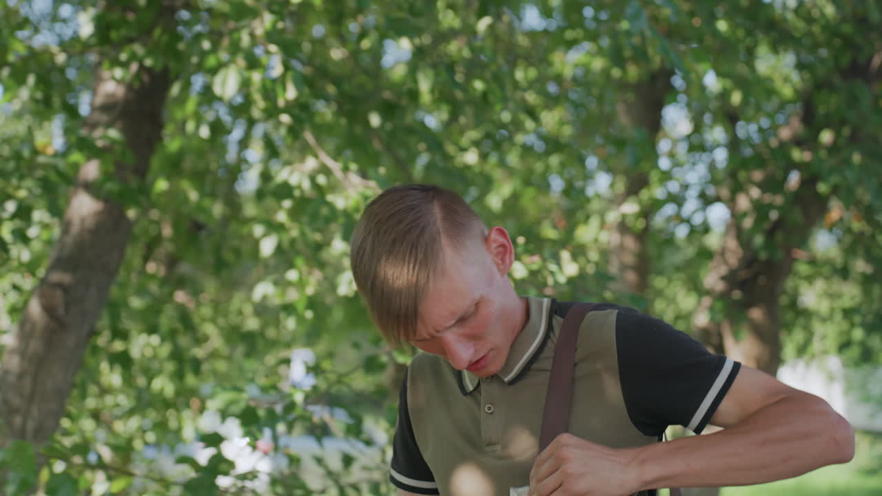 Young Man Alert In Park Scanning Surroundings And Adjusting Bag, Watchful Profile, Shifting Gaze To Street, Sunlight Flicker Through Leaves, Tense Shoulders, Commuter Vibe, Candid Reaction Closeup