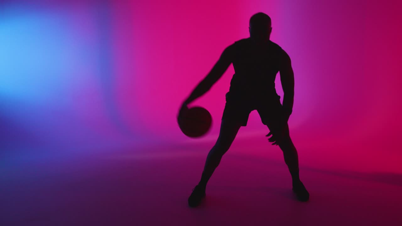 silueta de estudio de un jugador de baloncesto masculino driblando y rebotando la pelota contra un fondo iluminado de rosa y azul