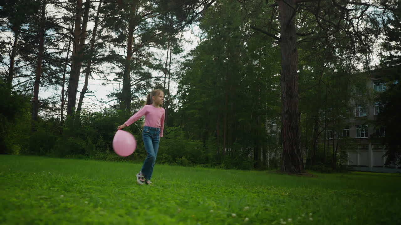 Girl in pink top and jeans walking across grassy field swinging pink balloon playfully while surrounded by tall trees, with a distant building partially visible through forested background