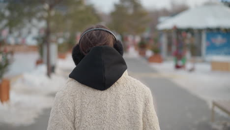 young girl in cozy beige winter jacket with black hoodie and fluffy earmuffs stares ahead before gently turning and looking up at sky, blurred background features snow-covered ground and trees