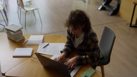 top view of a concentrated girl with curly hair sitting at a table and doing student work on a laptop in the library