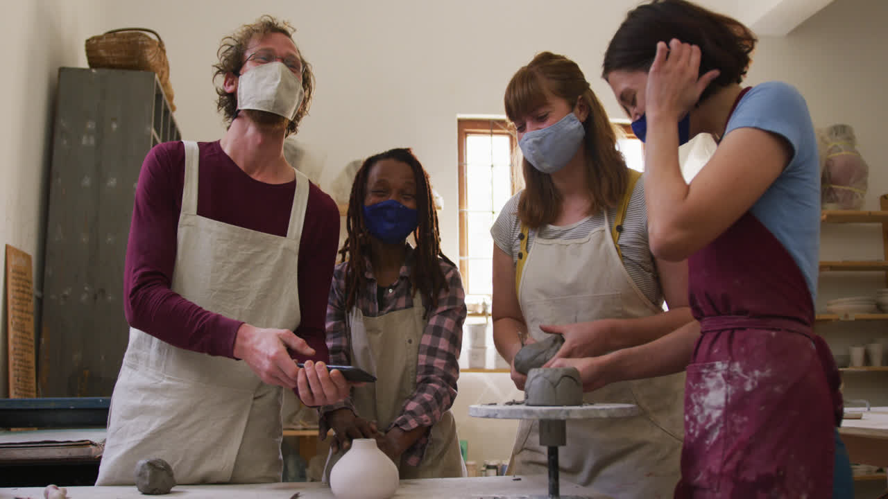 Diverse potters wearing face masks having a group video chat while creating pottery on potters wheel