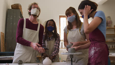 diverse potters wearing face masks having a group video chat while creating pottery on potters wheel