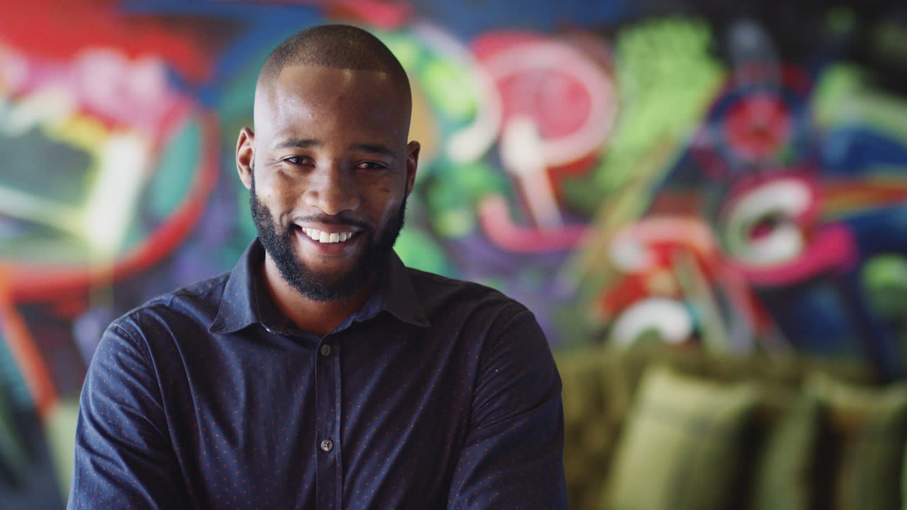 Jovem criativo negro milenar em frente a um mural no local de trabalho, sorrindo para a câmera, em um plano de cabeça e ombros.