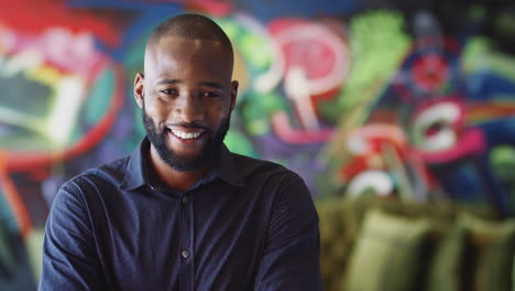 millennial black male creative in front of mural in workplace smiling to camera, head and shoulders
