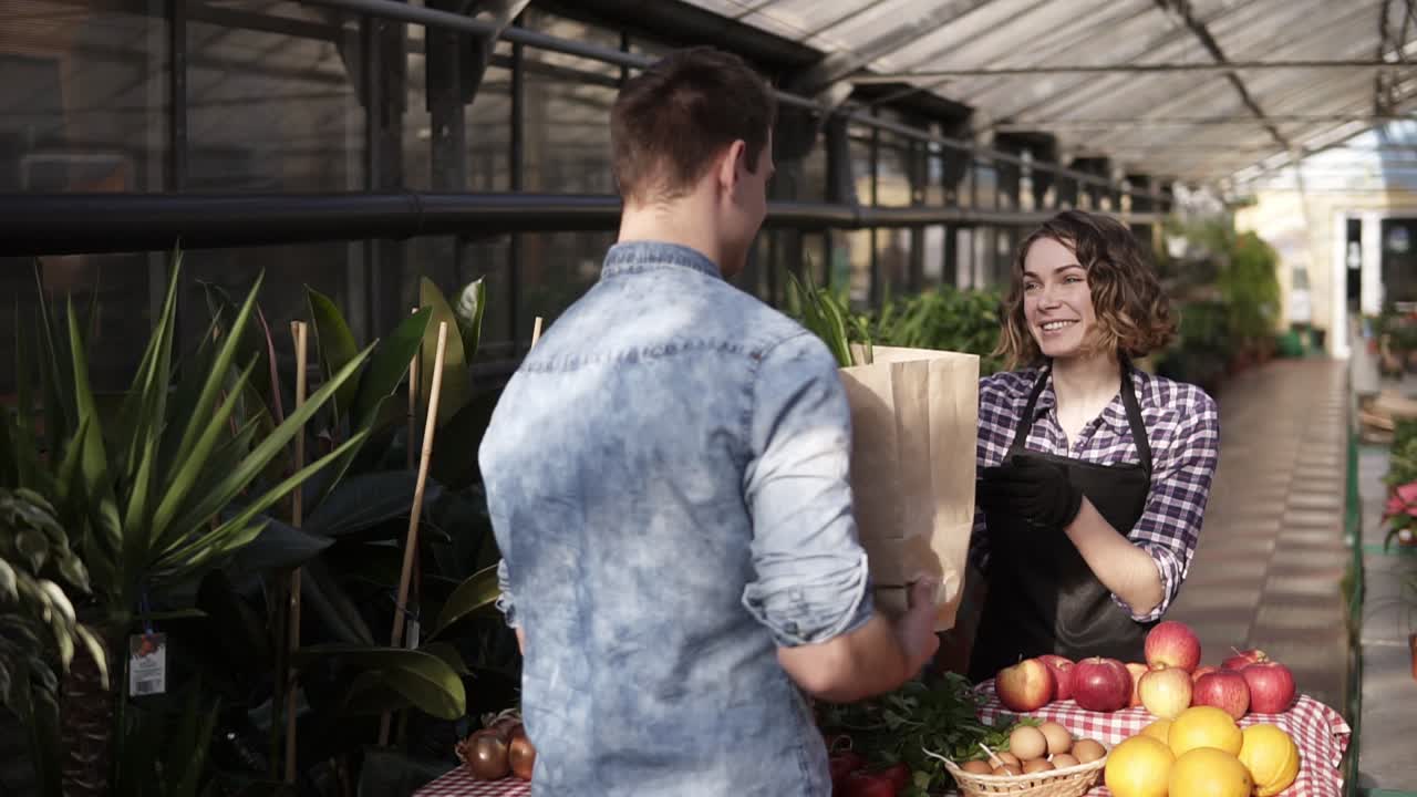 Cheerful saleswoman in black apron giving fresh vegetables in paper bag to customer in greenhouse market. Smiling woman selling vegetables to male customer. People and healthy lifestyle concept