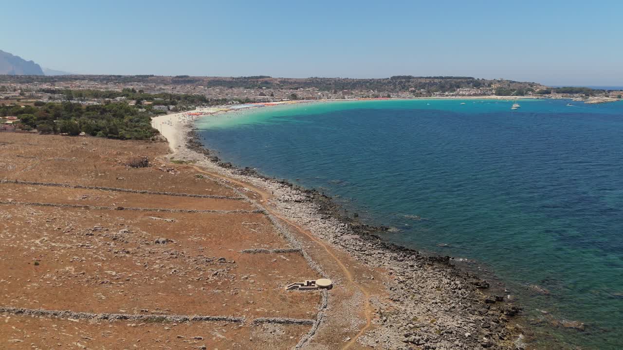 Zoom indietro aereo che rivela un bunker della Seconda Guerra Mondiale che si affaccia su una costa rocciosa siciliana e sul mare blu profondo, mostrando l'architettura militare storica in un paesaggio costiero