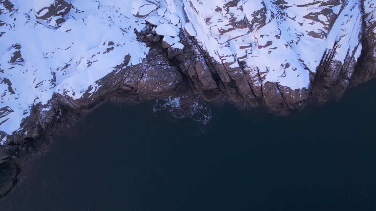 Vista aerea di una scogliera montuosa innevata, onde che si infrangono sulle rocce sottostanti, terreno accidentato, Isole Lofoten, Norvegia.