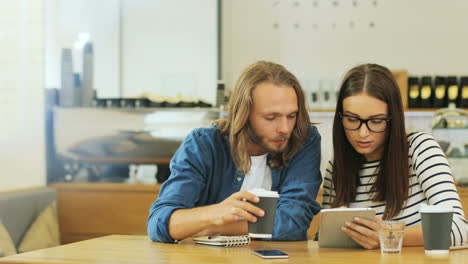 Caucasian-man-and-woman-friends-talking-and-watching-something-on-a-tablet-sitting-at-a-table-in-a-cafe