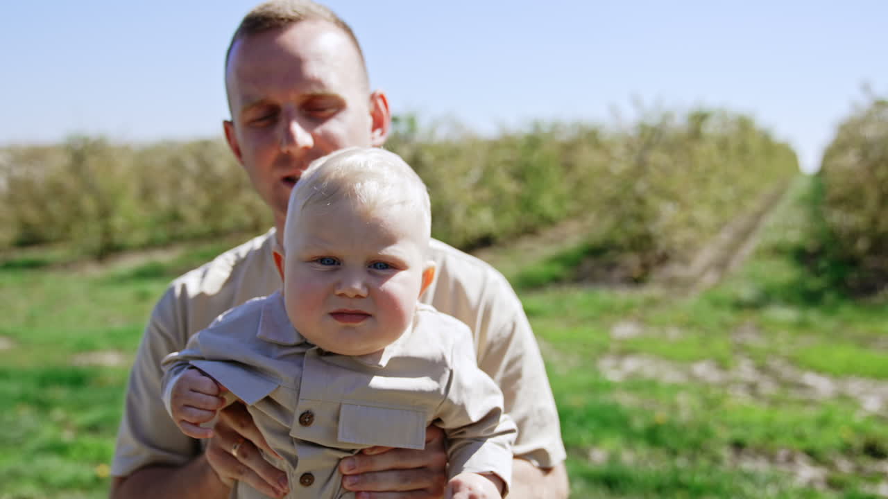 Caucasian man holding a cute blond baby boy in hands. Father shakes his son a little in front of camera. Young garden at backdrop.