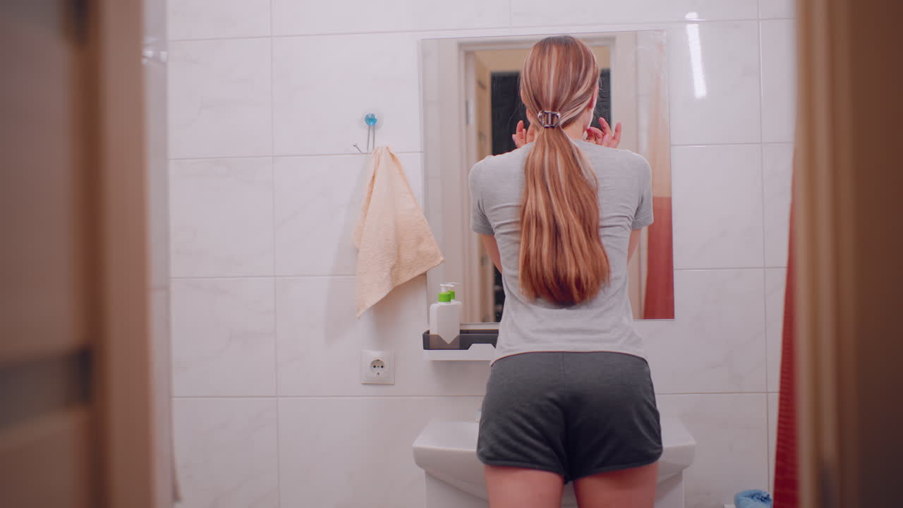 Back view of woman standing in bathroom looking at mirror, long hair clipped neatly behind with natural style, casual outfit, simple everyday routine moment in tiled interior