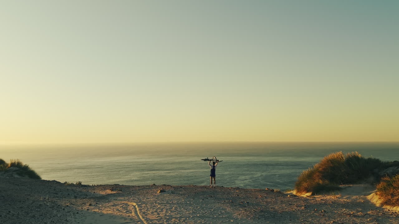 Person Holding Bicycle Above Head on Sandy Dune Overlooking Ocean at Sunset