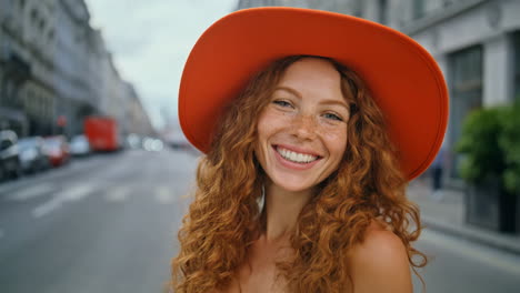 close-up portrait of a happy woman with red curly hair and an orange hat smiling on a city street