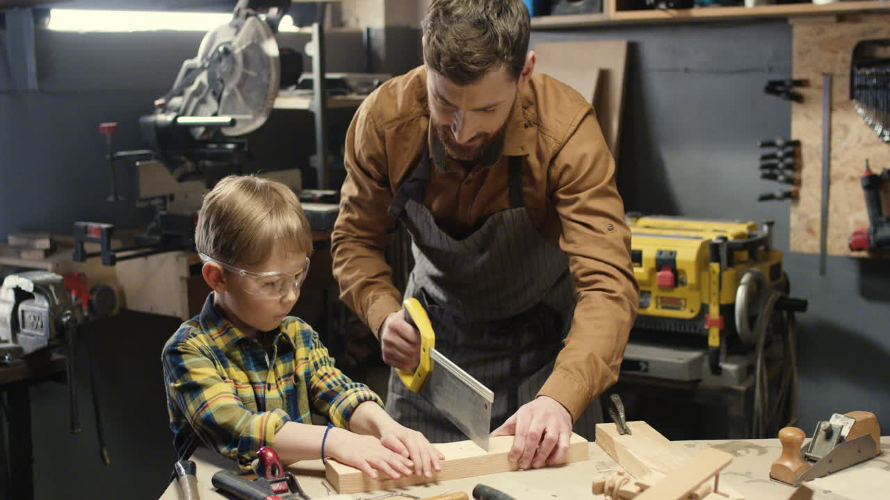 Caucasian carpenter man teaching his little son to work with hardwood and sawing timber in workshop