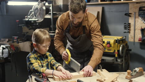 carpintero caucásico enseñando a su pequeño hijo a trabajar con madera dura y aserrando madera en el taller