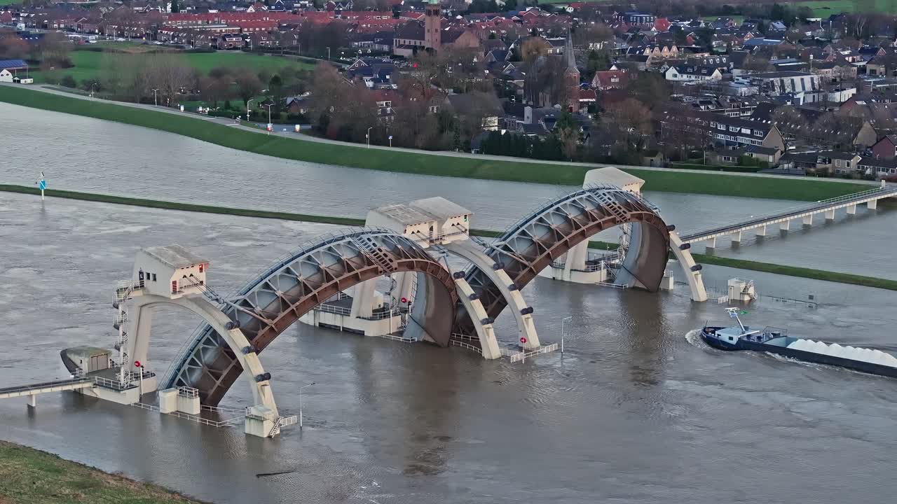 Zoomed in drone shot moving towards the weir of Driel with the town of Driel in the background during high water levels with the doors open and a cargo ship passing thru