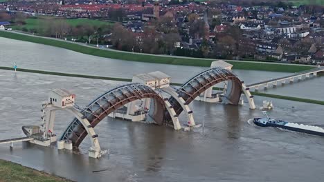 zoomed in drone shot moving towards the weir of driel with the town of driel in the background during high water levels with the doors open and a cargo ship passing thru