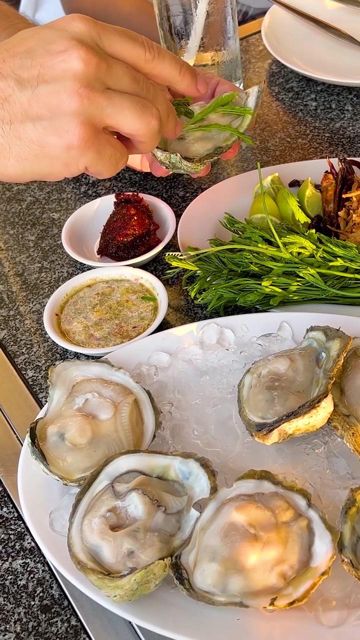 A vertical close-up shot of hands adding fresh greens and condiments to raw oysters.