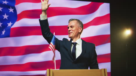 positive presidential candidate greeting audience pan around view of cheerful mature male politician in suit smiling and gesticulating while greeting audience under photo camera flashes against usa flag during election debate