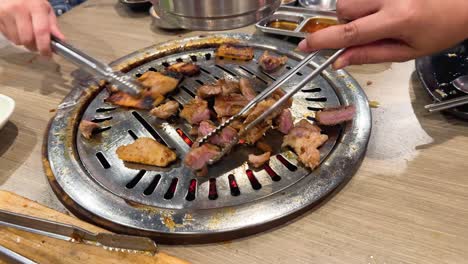 Hands grill pork and beef on a tabletop BBQ in a Bangkok restaurant. Warm lighting enhances the lively dining atmosphere