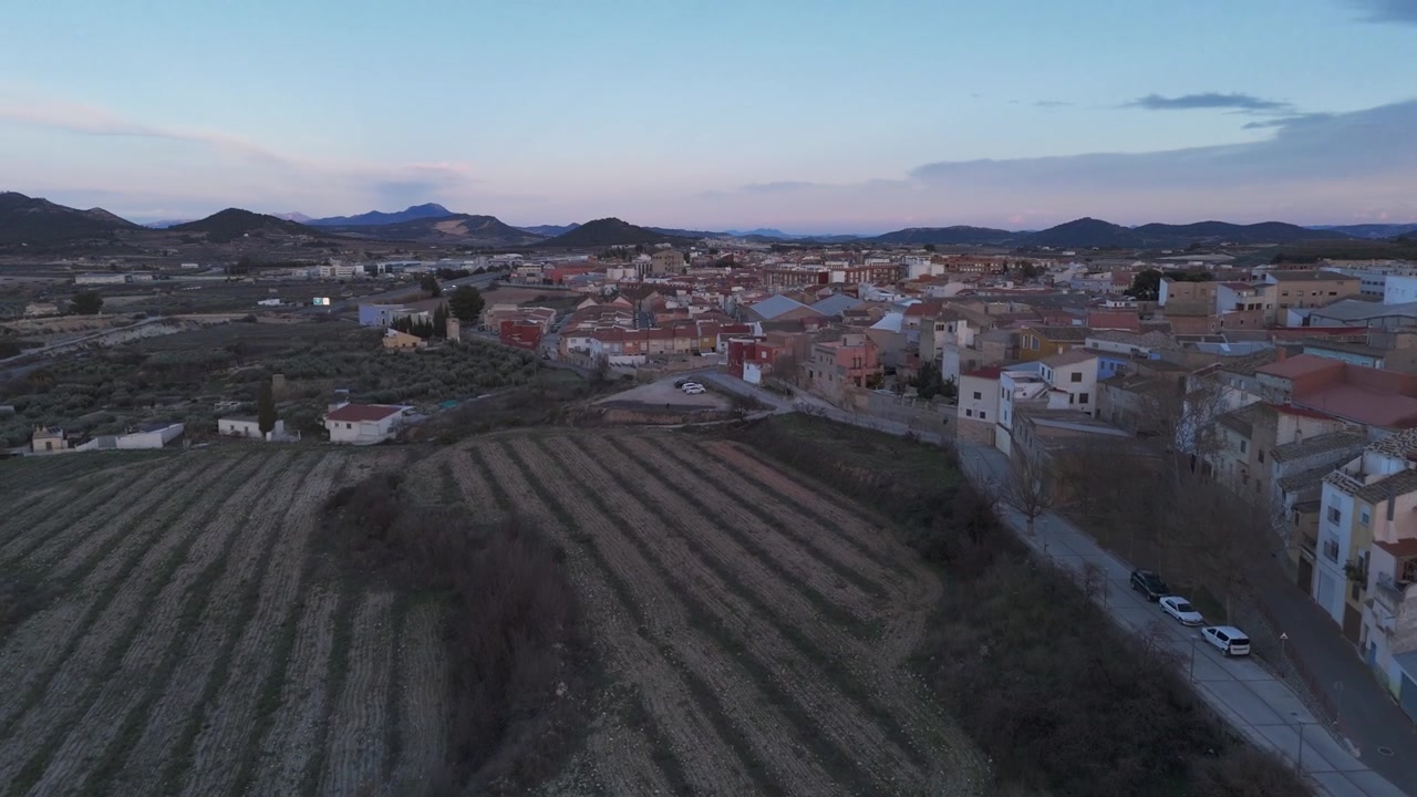 Timelapse con drone che vola sopra terreni agricoli rurali e vigneti verso la città di Bullas durante un tramonto colorato a Murcia, Spagna.