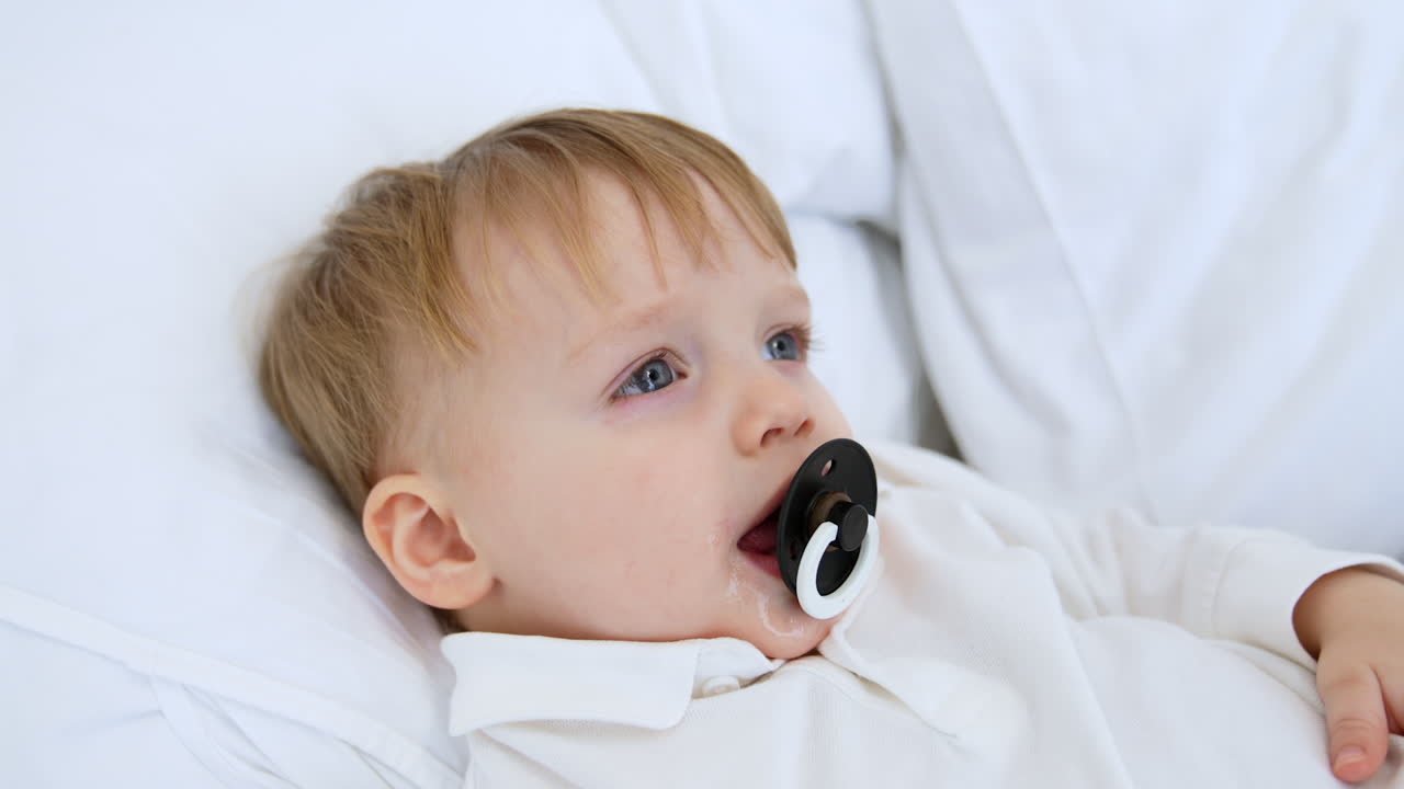 Cute happy Caucasian toddler boy in white sweater lies on the bed. Smiling kid with a pacifier in mouth. Close-up portrait.