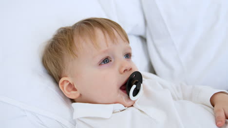cute happy caucasian toddler boy in white sweater lies on the bed. smiling kid with a pacifier in mouth. close-up portrait.