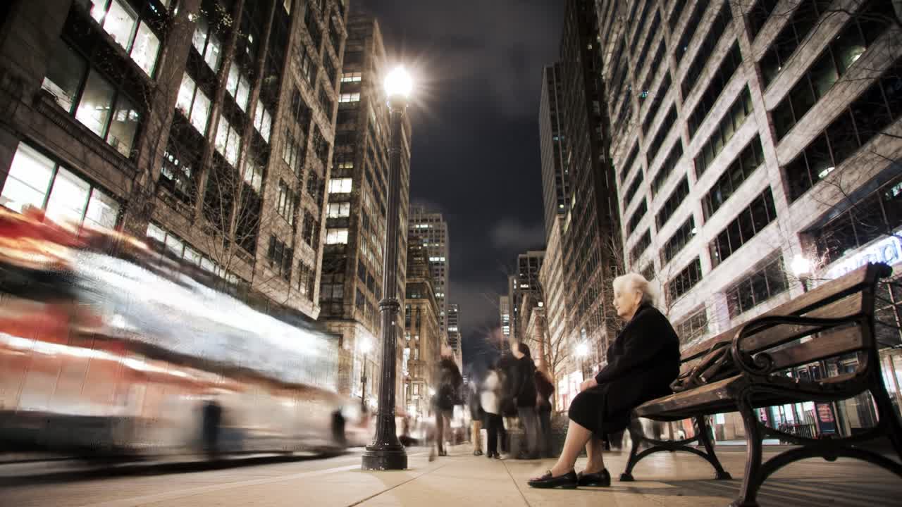 City streets come alive at night as a bus rushes by while a woman relaxes on a bench. Pedestrians stroll, enjoying the vibrant urban ambiance and illuminated buildings.