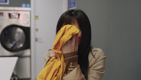 With-bangs-framing-her-face,-an-Asian-woman-takes-a-moment-to-breathe-in-the-fresh-scent-of-her-recently-laundered-clothes-at-the-laundromat,-wearing-a-content-expression